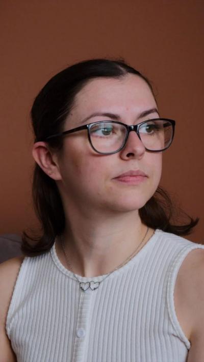 A woman with brown hair and dark rimmed glasses wearing a white vest top looks to her left. Behind her is an orange wall