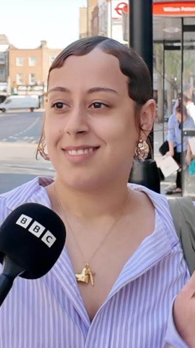 Person speaking into a BBC microphone on a sunny urban street with shops and a bus stop in the background.