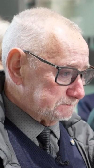 Geoff Burrow in a grey shirt and tie with a blue jumper, grey coat and dark glasses has short grey hair and a goatee.