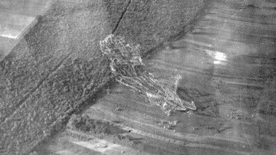A black and white aerial photo of the skeleton of a large airship. It is in a field, with the nose of the skeleton in a densely wooded area, and the body and tail on a grassy field. There are people in the field.