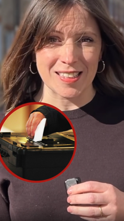 A reporter looks at the camera while talking. She is wearing a brown t-shirt and has short brown hair. There is a circular image of a person placing a ballot paper into a ballot box inset in the picture. 