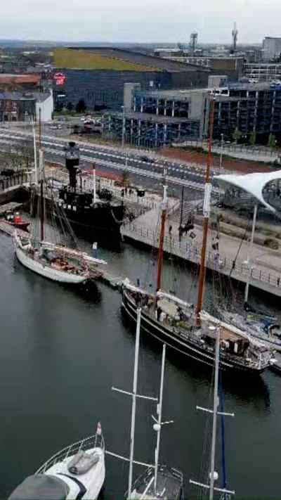 An aerial view shows a marina with white yachts and two tall ships moored at Hull Marina. Modern buildings, a road, and the white Murdoch's Connection bridge line the water’s edge in the background.