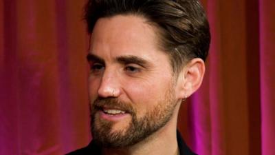 Man with brown hair and beard sat in front of red curtain.