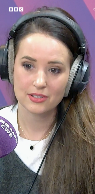 Woman sitting in a radio studio with headphones on, brown hair, red lipstick. Talking into a purple microphone and there's a purple background to the picture.