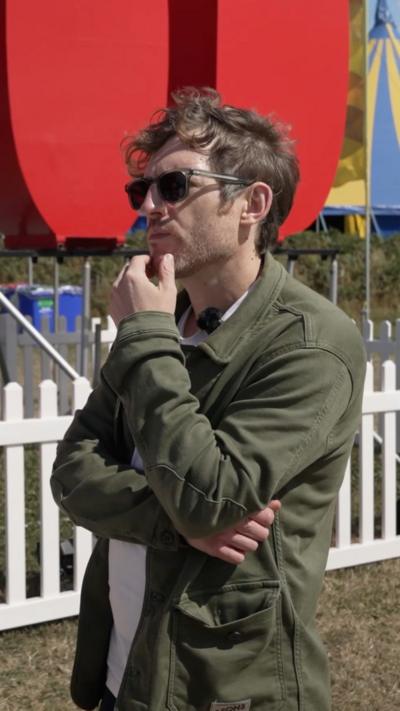 Elis James, the comedian, in sunglasses stood outside with his hand on his chin in a thinking pose