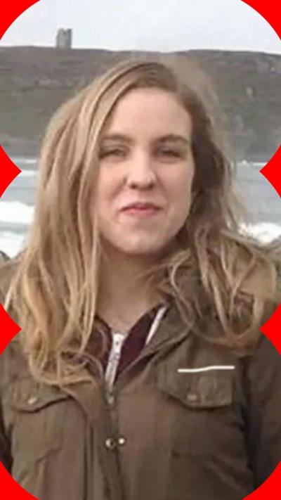 A woman with long blonde hair is stands outside in front of the sea. She is wearing a brown coat with a maroon zip top underneath.