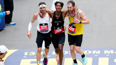 Three men wearing black shorts and different coloured running vests jog towards the finish line of the Boston Marathon with their arms around each other. Robson De Oliveira, who has short, dark hair and a moustasch and is wearing a white vest and a white headband. Ajay Haridasse has short, dark hair and a black vest. Aaron Beggs has short brown hair and a beard and is wearing a yellow vest with a blue stripe.