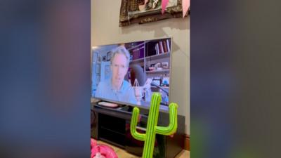 A large flat-screen TV can be seen in front of a cream or white wall atop a black TV stand. Rob Brydon the actor that plays Uncle Bryn in the sitcom Gavin and Stacey is on the television looking directly into the camera. There is also an inflatable cactus and pink bunting hanging above the walls and large horizontal picture frame above the television, the painting inside is not visible. 