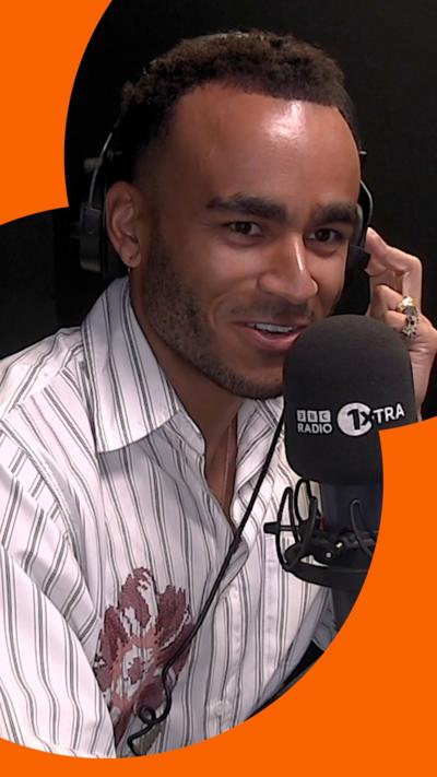 Munya Chawawa smiles, wearing a stripy shirt in front of a R1Xtra microphone.