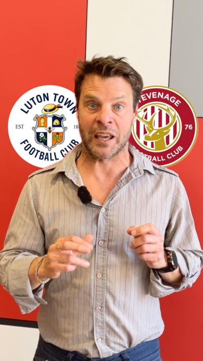 BBC presenter stands in front of screen with club badges for Luton Town and Stevenage FC on it. He is wearing a beige shirt and blue trousers. 
