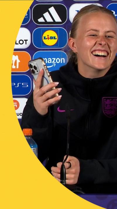 Hannah Hampton sat behind a table during a press conference laughing as she holds up her mobile phone