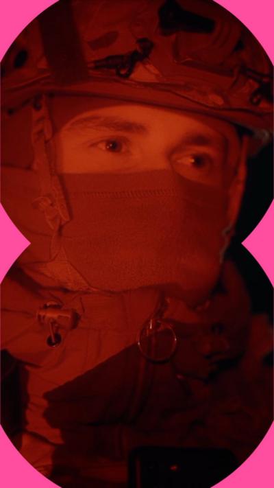 The face of a man wearing a balaclava and an army helmet, who is lit up in red light at night.