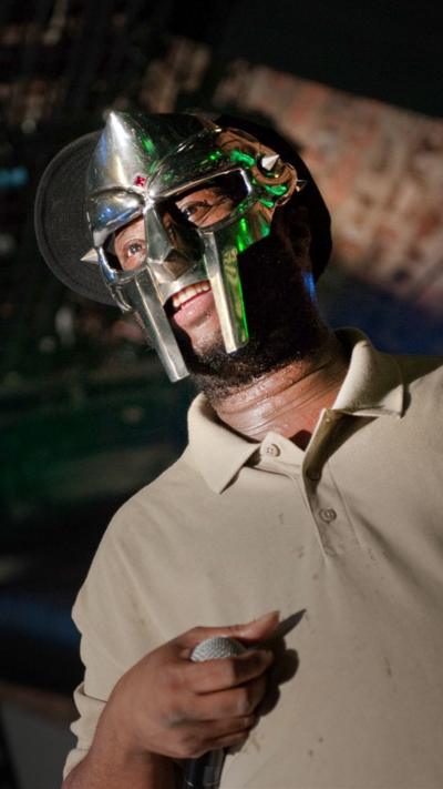 MF Doom wearing a silver face mask and smiling. He is holding a microphone in his right hand