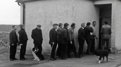 Black and white image of a line of men queuing outside a rural public toilet.