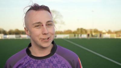 Person with face piercings wearing a purple top outdoors on artificial, green playing pitch
