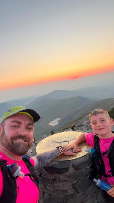 A man and a boy at the top of Yr Wyddfa touching the top of the peak as the sun rises. They are both in pink t-shirts