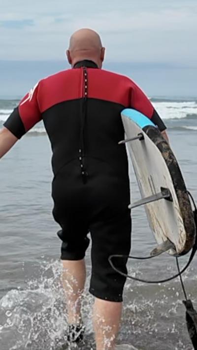 A man in a red and black wetsuit carrying a surfboard walking away from the camera into the sea