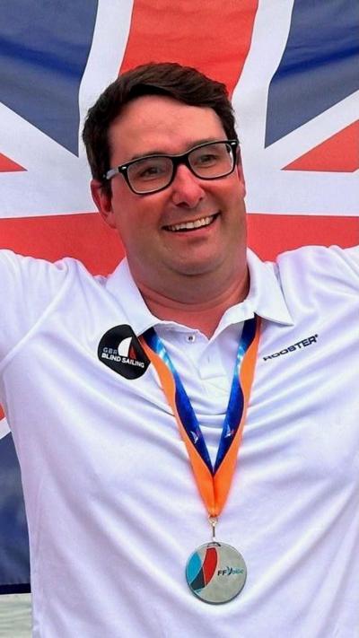 A man with brown hair and black glasses stood in front of a union flag. he's wearing a white shirt and had a gold medal around his neck. The ribbon holding the medal is blue and orange
