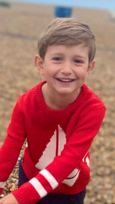 Young boy, Benedict, wearing a bright red jumper looks towards the camera outdoors, with a softly blurred background creating a natural portrait.