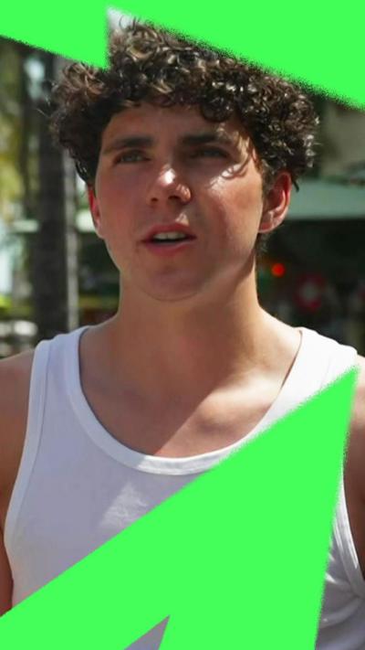 A young man with curly dark hair, wearing a vest and speaking to a BBC reporter