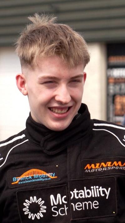 Ally Chalmers, a 17-year-old boy with short, blond hair. He is looking off to the side and is smiling. He is wearing a black race suit with the names of sponsors on it. 