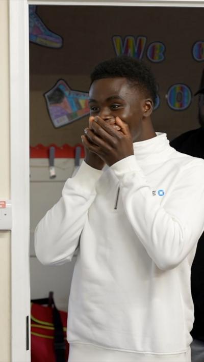 Bukayo Saka in a white zip-up shirt stands nervously, with hands covering his mouth, in a classroom setting.