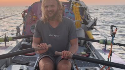 Man with long hair sits in a rowing boat, behind him is equipment, dusk is falling over the Pacific Ocean