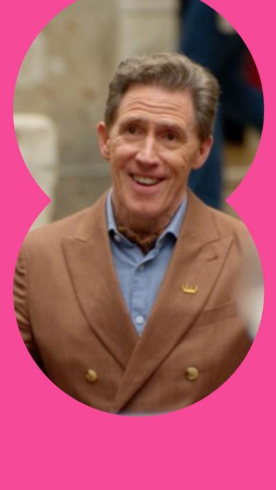 Rob Brydon opens his arms while singing to a table of people. He wears a brown suit.