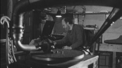 A man sits at a tape recorder. A record player is in the foreground of the image