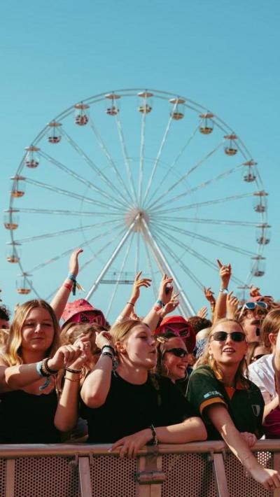 A crowd of festivalgoers stands at the front barrier with arms raised while a large Ferris wheel rises in the background against a clear blue sky.