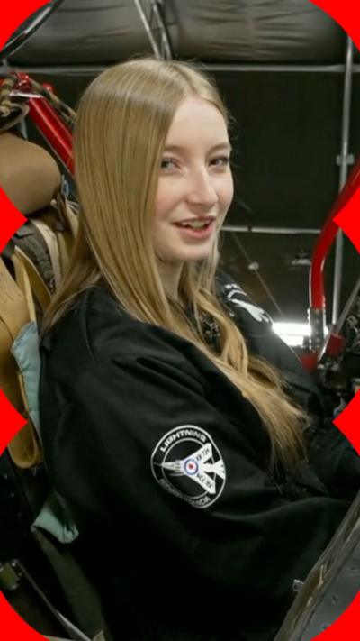 Grace sits in the fighter jet and looks at the camera. She is wearing a black boiler suit. 