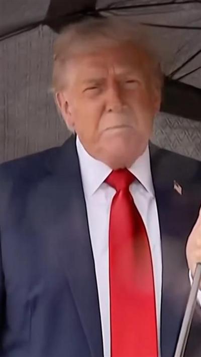 Donald Trump, wearing a suit, stands beneath an umbrella