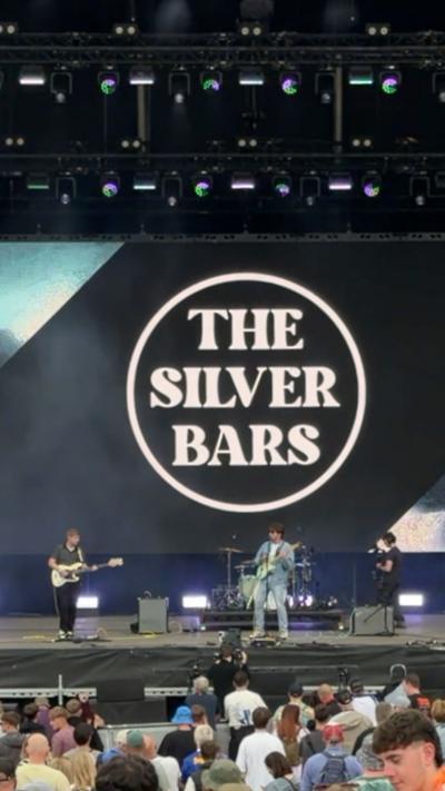 The Silver Bars perform on the main stage at Kendal Calling in front of a large crowd. A giant screen behind the band displays The Silver Bars logo in bold white lettering. Audience members watch from the front of the stage as the band plays under the festival lighting.