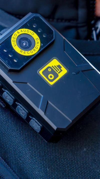 A close up of a body worn camera clipped onto the collar of a jacket. The camera is black with yellow logos.