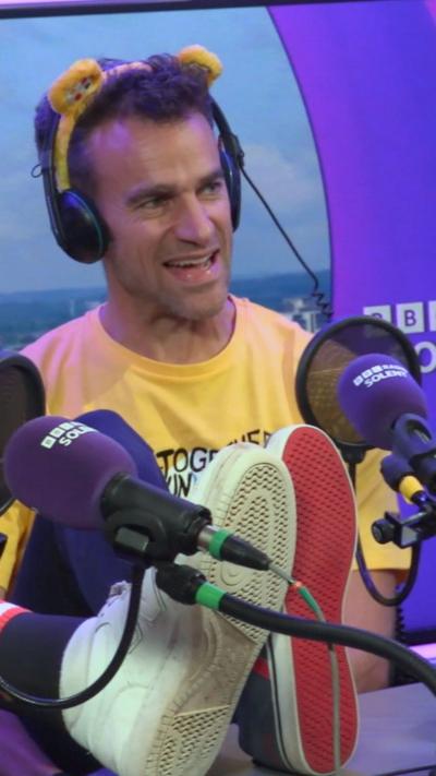 Steve Harris has short brown hair and wears Pudsey ears and headphones with a yellow top sitting in the radio studio against a purple backdrop