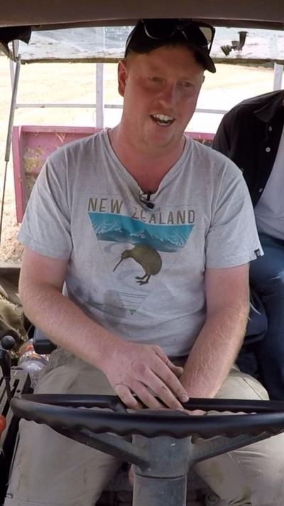 A farmer in a pale t-shirt with New Zealand on the front and a Kiwi bird. He's wearing a black cap and driving a tractor