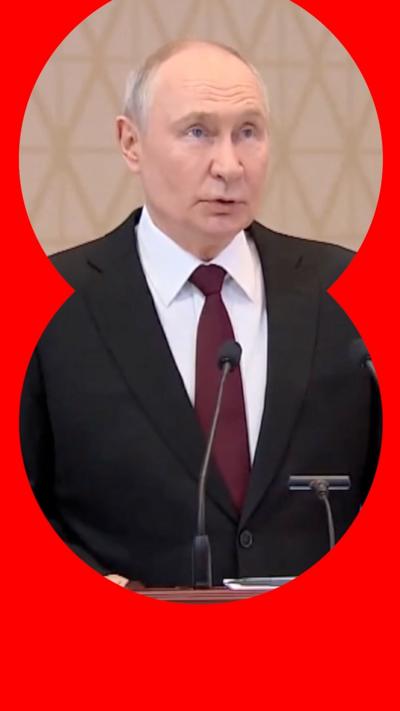 Vladimir Putin has short hair and wears a black suit as he stands at a podium and speaks into a microphone
