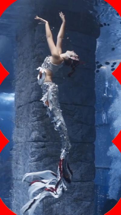 A woman in a mermaid costume reaches her arms upwards underwater, with a male statue in the foreground.