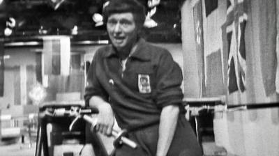 Black and white image of Blue Peter presenter John Noakes holding a small dumbbell and looking out of breath. 