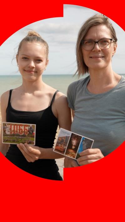 A teenage girl and a woman stand on a beach, holding postcards, looking to camera.