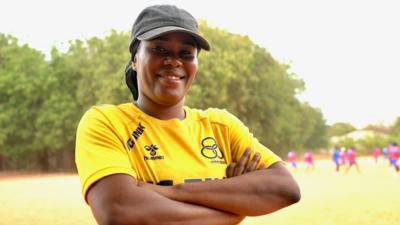 Sainey Mboge seen from waist up on a sandy pitch looking into the camera and smiling with arms folded. She wears a yellow training top with various black logos on it and a grey cap. In the background, blurred, are seen a group of young girls playing football in blue kit, with some wearing purple bibs.
