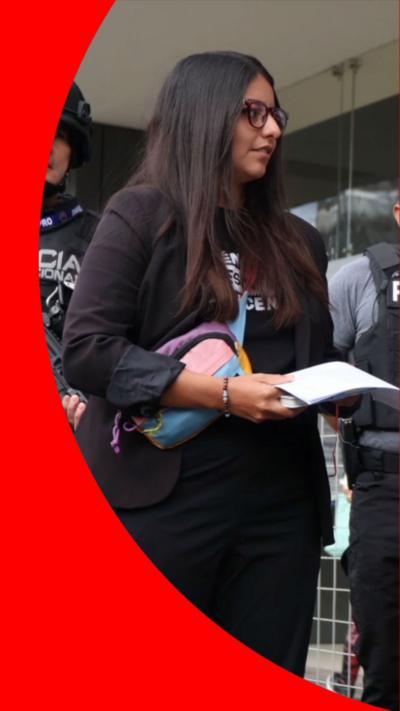 A woman with long dark hair wearing glasses and dressed in black, holds a piece of white paper.