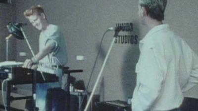 Two members of band, Depeche Mode, stand at electronic instruments in a rehearsal studio. 