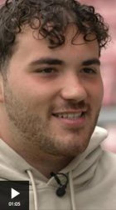 A close up shot of a man smiling. He has dark curly hair and a short beard, and is wearing a cream coloured hoodie