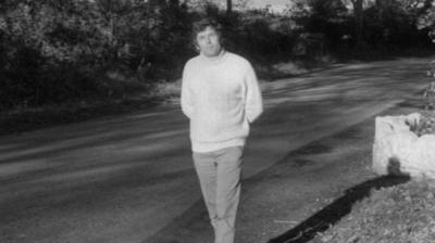 A reporter in a thick woollen jumper stands on the fairy bridge of the Isle of Man.