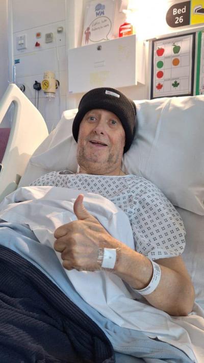 A man in a hospital bed wearing a black bobble hat giving the thumbs-up sign