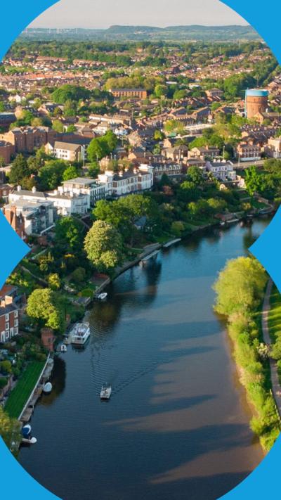 An aerial view of Chester in the sunshine - there is a river lined with trees and brown buildings in the background