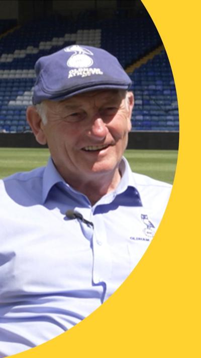 Frank Rothwell is wearing a light blue shirt and a navy flat cap. He is sat next to the Oldham Athletic pitch. 