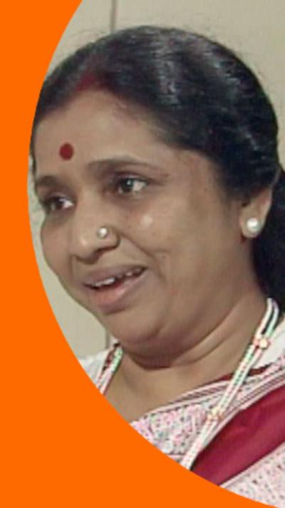 A woman, Asha Bhosle, with dark hair smiling. She is wearing a white, red and pink top.