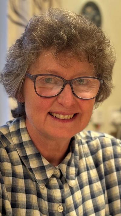 A woman in a yellow blue check shirt sat in a kitchen. She has grey hair and black glasses